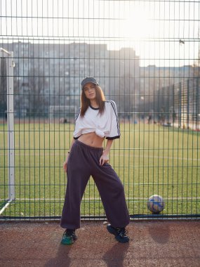 Güneş batarken tel örgülerin yanında duran beyaz tişörtlü, eşofman altı ve beyzbol şapkalı güzel bir kız. Açık hava futbol sahasında, topun yanında ince beli olan bir kadın model. Şehir modası.
