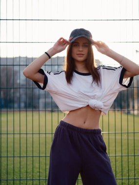 Güneş batarken tel örgülerin yanında durup kameraya bakan beyaz tişörtlü ve beyzbol şapkalı güzel bir kız. Açık hava futbol sahasında ince beli olan bir kadın model. Şehir modası.