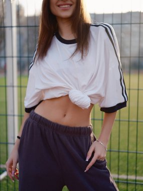 Gün batımında tel örgülerin yanında duran beyaz tişörtlü güzel bir kız. Açık hava futbol sahasında ince beli olan bir kadın model. Şehir modası. Vücut parçası görünür.