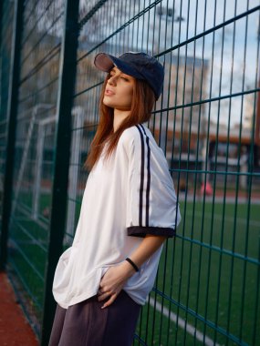 Futbol sahasında duran çekici bir model. Tel örgü çitlere ve yeşil çimlere karşı poz veren modaya uygun tişört ve şapkalı kız profili. Kentsel yaşam tarzı, gençlik modası ve spor estetiği..