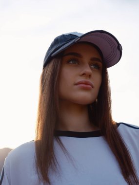 Gün batımında gökyüzüne karşı poz veren çekici bir model. Moda şapkalı ve beyaz tişörtlü bir kız kendinden emin bir ifadeyle mesafeye bakıyor. Şehir güzelliği ve gençlik modası. Düşük açı.