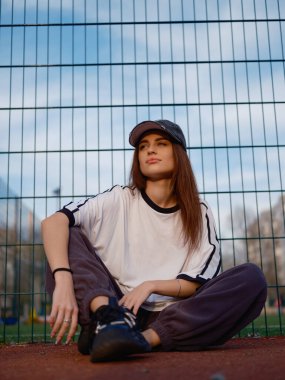 Kırmızı lastik kortta tel örgülere ve mavi gökyüzüne karşı oturan çekici bir model. Beyaz tişörtlü ve beysbol şapkalı bir kız rahat poz veriyor. Şehir modası ve gençlik hayatı. Düşük açı.
