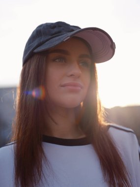 Gün batımında gülümseyen çekici mankenin yakın çekimi. Beyaz tişörtlü ve kepli bir kız sıcak güneş ışığı ve renkli bir mercekle dışarıda poz veriyor. Şehir güzelliği, gençlik ve dinginlik kavramı.