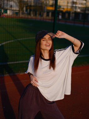 Gözleri kapalı, gülen ve şapkasına dokunan güzel bir kızın samimi portresi. Gün batımında kırmızı bir açık hava spor sahasında poz vermek. Şehir tarzı ve pozitif titreşimler.