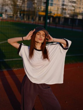 Açık hava atletizm sahasında gözleri kapalı öpüşen çekici bir manken. Beyaz tişörtlü ve eşofmanlı kız günbatımının tadını çıkarıyor. Gençlik modası, mutluluk ve şehir yaşam tarzı kavramı.