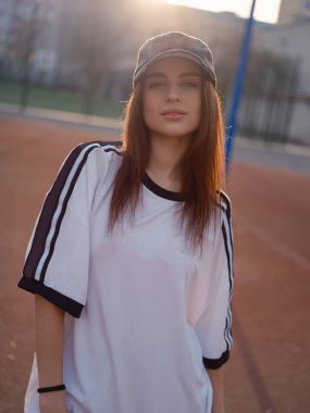 Kameraya tarafsız bir ifadeyle bakan bir kadın modelin yakın plan portresi. Sıcak güneş ışığı, açık hava spor sahasında koyu renk saçlarının ve beyzbol şapkasının etrafında bir parıltı yaratır..