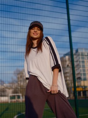 Büyük boy tişört ve beysbol şapkalı, gülümseyen kadın modelin düşük açılı görüntüsü. Açık havada, arka planda mavi gökyüzü ve yerleşim binaları olan bir spor alanında poz veriyor..