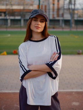 Atletizm sahasında duran beyaz tişört ve beysbol şapkası giyen bir manken. Gün batımında yerleşim binaları ve yeşil çimenlikli şehir arkaplanı.