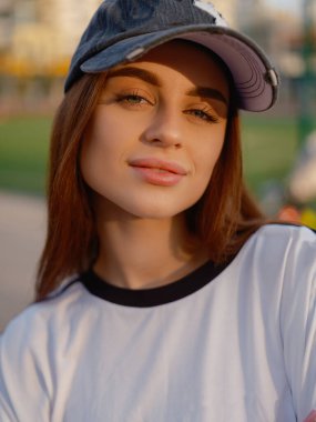 Büyük boy beyaz tişört giyen güzel bir kız ve siyah şapkalı yumuşak bir gülümsemeyle kameraya bakıyor. Güneş ışığı, gün batımında açık hava spor sahasında yüzünü ve gözlerini ön plana çıkarıyor..