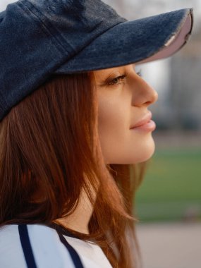 Çekici bir kızın huzurlu bir ifadeyle bakışlarının detaylı portresi. Sıcak altın saat güneş ışığı teninde ve kirpiklerinde yumuşak bir parıltı yaratıyor. Kentsel spor sahası geçmişi.