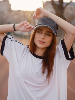 Güzel bir kız açık hava spor sahasında elleri şapkasının yanında poz veriyor. Yumuşak altın saat ışığı ve sıcak arka plan huzurlu bir şehir hayatı ortamı yaratır..