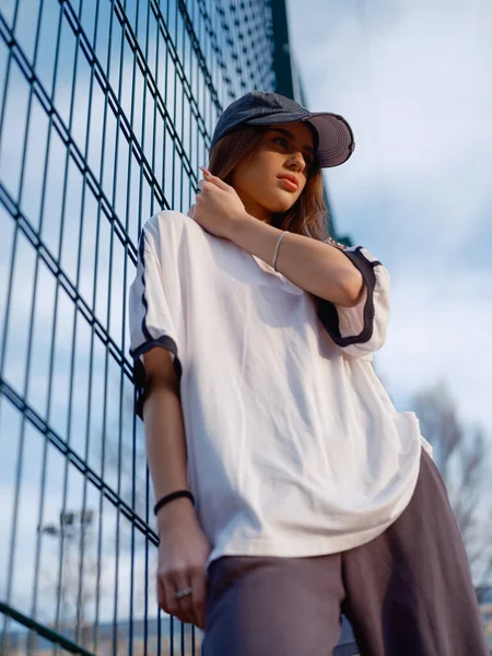 Beyaz tişörtlü ve kapaklı çekici bir kadın manken spor sahasının tel örgüsüne yaslanıyor. Arka planda gökyüzü olan güzel bir kız. Sokak modası ve gençlik hayatı.