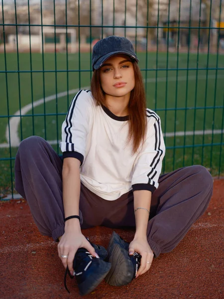 Açık hava spor sahasının kırmızı kauçuk yüzeyinde rahat poz veren çekici bir model. Beyaz tişörtlü ve kepli bir kız tel örgülere ve futbol sahasına karşı. Şehir modası ve gençlik yaşam tarzı.