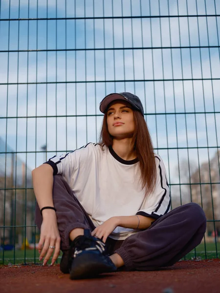 Kırmızı lastik kortta tel örgülere ve mavi gökyüzüne karşı oturan çekici bir model. Beyaz tişörtlü ve beysbol şapkalı bir kız rahat poz veriyor. Şehir modası ve gençlik hayatı. Düşük açı.