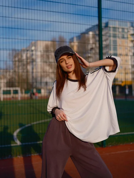 Güneşli bir günde tel örgülerin yanında duran beyzbol şapkalı ve spor giysili bir kadın model. Arka planda yeşil bir futbol sahası ve konut binaları yer alıyor..