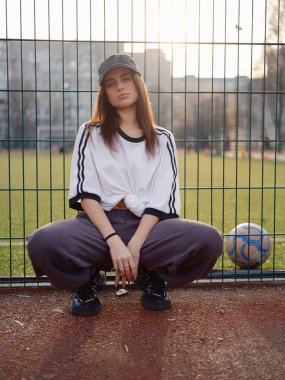 Beyaz tişörtlü ve beyzbol şapkalı çekici bir kız açık hava futbol sahasında çömelmiş poz veriyor. Gün batımında çenesinin altında eli olan bir kadın model. Şehir modası, gençlik yaşam tarzı ve spor estetiği.