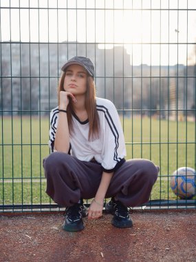 Beyaz tişörtlü ve beyzbol şapkalı çekici bir kız açık hava futbol sahasında çömelmiş poz veriyor. Gün batımında çenesinin altında eli olan bir kadın model. Şehir modası, gençlik yaşam tarzı ve spor estetiği.