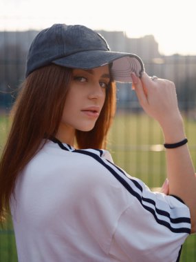 Omzunun üzerinden kameraya bakan çekici bir kadın model. Gün batımında beyaz tişörtlü ve şapkalı bir kız spor çitlerine karşı poz veriyor. Şehir modası ve güzellik kavramı.