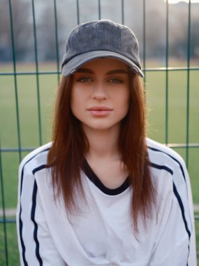 Çekici kadın modelin portresi kameraya bakarken dalgalanıyor. Gün batımında beyaz tişörtlü ve şapkalı bir kız spor çitlerine karşı poz veriyor. Şehir modası ve güzellik kavramı.