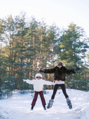 Neşeli anne ve kızı, karlı kış ormanlarında birlikte zıplıyorlar. Slav tarzı lüks kürk şapkalar ve paltolar giyen bir aile. Açık havada aktif kış tatili eğlencesi. Güneşli bir gün, çam ağaçları, kar.