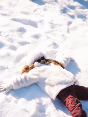 Lüks kürk şapkalı ve güneş gözlüklü neşeli çocuk taze karın üzerinde yatıyor. Güneşli bir kış ormanında açık havada oynayan bir kız. Slav modası ve çocukluk eğlencesi. Aktif boş zaman. Yüksek açı görünümü.