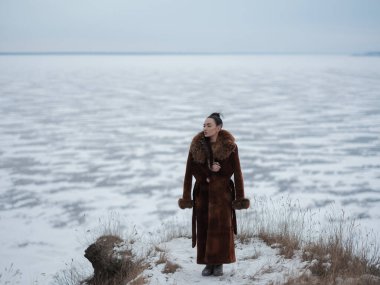 Yüksek tepelerde uzun kürklü, zarif genç bir kadın. Donmuş deniz altyapısı olan manzaralı kış manzarası. Arktik doğaya bakan şık, sıcak giysiler içinde bir model..