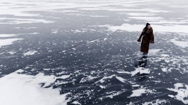 Lüks kahverengi kürk giyen genç bir kadın donmuş su yüzeyinde yürüyor. Beyaz kar desenli koyu renkli buzun yüksek açılı görüntüsü. Minimalist kutup buzlu doğa manzarası. Boşluğu kopyala.