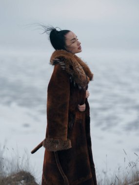 Kapalı gözlerle poz veren güzel genç bir kadının yakın çekimi. Büyük yakalı kahverengi kürk manto giyiyor. Kış atmosferi bulanık arkaplan ve yumuşak gün ışığıyla.