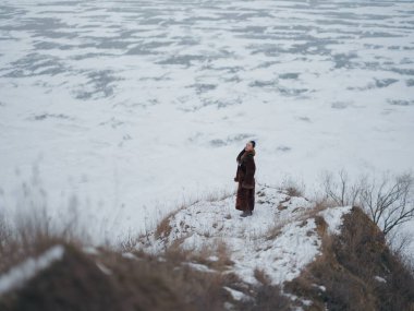 Uçurumun kenarına poz veren zarif bir kadının yüksek açılı çekimi. Arka planda buz desenleri olan engin donmuş deniz. Lüks kürk giyen kadın modelle minimalist kış manzarası.