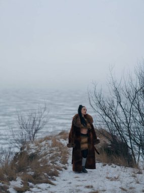 Kışın açık havada poz veren kahverengi kürklü ve uzun paltolu genç kadın donmuş göle ve sisli ufka karşı çıplak ağaçlarla kaplı tepede. Arktik moda portresi vahşi doğada.