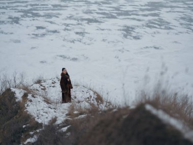 Uçurumun kenarına poz veren zarif bir kadının yüksek açılı çekimi. Arka planda buz desenleri olan engin donmuş deniz. Lüks kürk giyen kadın modelle minimalist kış manzarası.