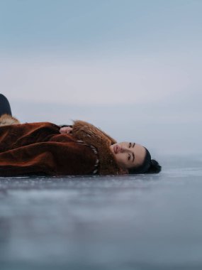 Kışın buz yüzeyinde uzanan güzel genç bir kadının yakın çekimi. Büyük yakalı, kahverengi kürk giyen bir manken. Artistik arktik portre. Önü bulanık ve gün ışığı yumuşak..