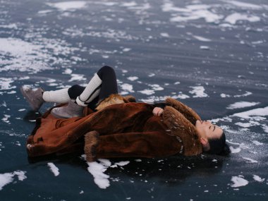 Lüks kışlık kürklü genç bir kadın ve donmuş su yüzeyinde uzanan bacak ısıtıcıları. Kontrastlı arka planda beyaz kar lekeleri olan koyu renk buz. Kuzey Kutbu 'nda poz veren modaya uygun bir kadın modeli..