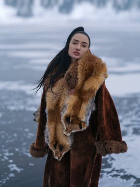 Lüks kış kıyafetleriyle dışarıda poz veren güzel genç bir kadın. Uzun kürklü manken ve kafalı egzotik kırmızı tilki kürkü. Yumuşak gün ışığı ve kutup atmosferiyle donuk göl arka planı.