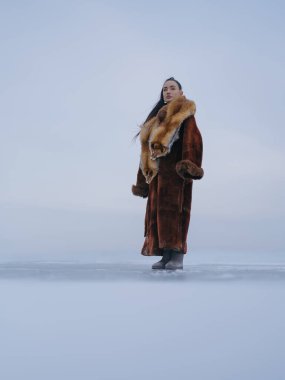 Lüks kahverengi kürklü, kırmızı kürklü, kafası donmuş gölde kışın gökyüzüne karşı poz veren zarif bir kadının alçak açılı görüntüsü. Soğuk minimalist geçmişe sahip modaya uygun bir arktik portre..