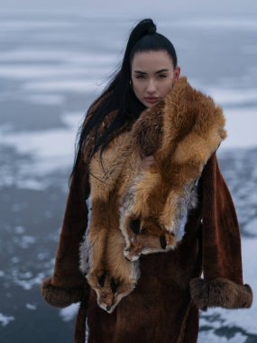 Lüks kış kıyafetleriyle dışarıda poz veren güzel genç bir kadın. Uzun kürklü manken ve kafalı egzotik kırmızı tilki kürkü. Yumuşak gün ışığı ve kutup atmosferiyle donuk göl arka planı.