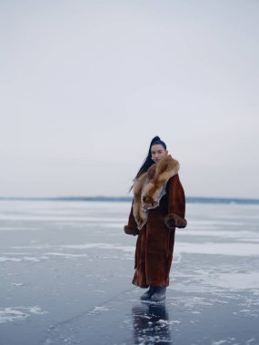 Engin donmuş su yüzeyinde lüks kış kıyafetleri içinde poz veren zarif genç bir kadın. Uzun kürklü manken ve kafalı kırmızı tilki kürkü. Kış kutup doğası ve bulutlu gökyüzü.