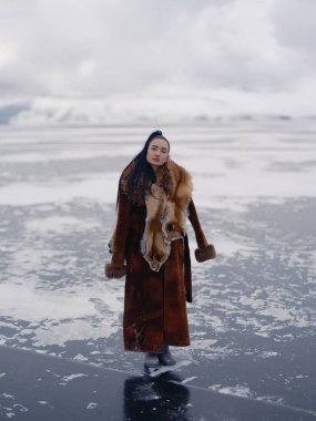 Engin donmuş su yüzeyinde lüks kış kıyafetleri içinde poz veren zarif genç bir kadın. Uzun kürklü manken ve kafalı kırmızı tilki kürkü. Kış kutup doğası ve bulutlu gökyüzü.