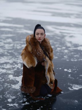 Uzun kahverengi kürklü ve egzotik tilki kürklü güzel bir kadın dizlerinin üstünde poz veriyor. Karla kaplı donmuş su yüzeyine yakın çekim. Soğuk atmosferli arktik moda portresi.