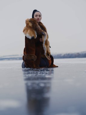 Lüks kürklü ve tilki kürklü zarif genç kadın kameraya bakarak dizlerinin üstünde poz veriyor. Düşük açılı çekim, donmuş gölün yansıması. Kuzey Kutbu doğasında sisli bir moda portresi.