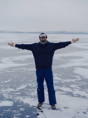 Kayak gözlüklü ve kışlıklı neşeli genç adam, donmuş su yüzeyinde kollarını açmış, koyu buz üzerinde kar desenleri olan. Özgürlük kavramı, seyahat, Kuzey Kutbu doğasında kış macerası.