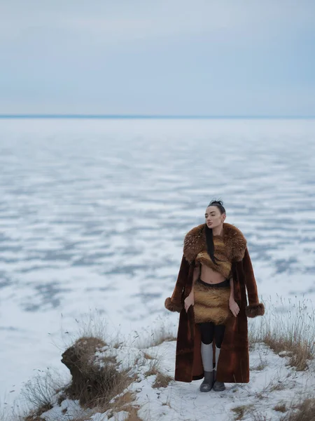 Donmuş göle bakan karlı sahilde, kısa kürklü bluz ve etekli genç bir kadın. Kutup manzarasında kış modası portresi, lüks kıyafetler ve çıplak cilt kontrastı..