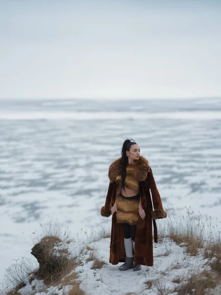 Donmuş göle bakan karlı sahilde, kısa kürklü bluz ve etekli genç bir kadın. Kutup manzarasında kış modası portresi, lüks kıyafetler ve çıplak cilt kontrastı..
