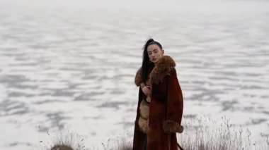 Zengin kahverengi kürklü şık bir kadın arka planda donmuş gölün kenarında uçurumun kenarında poz veriyor. Soğuk ve bulutlu bir günde zarafet, kış lüksü ve üst düzey moda..
