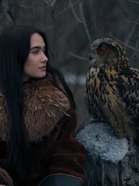 Kürk mantolu genç bir kadın Avrasya kartal baykuşuna bakıyor. Kuş, kışın ağaçlık alanda ağaç kütüğüne tünemiş. Karamsar atmosferde insan ve vahşi yaşam arasındaki duygusal bağ.