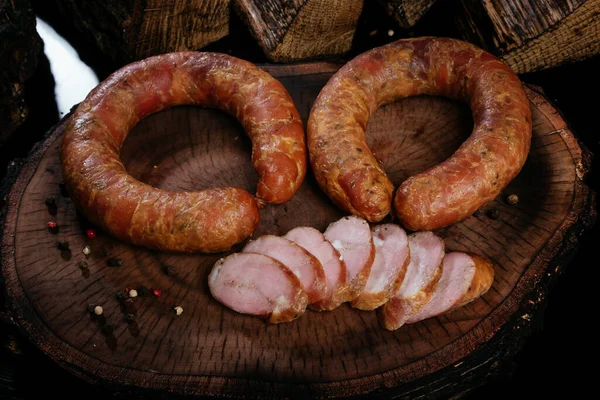 Zwei Ringe Räucherwurst auf einem Brett, auf einem Hintergrund aus verbranntem Brennholz. Mehrere Wurststücke in Scheiben geschnitten. — Stockfoto