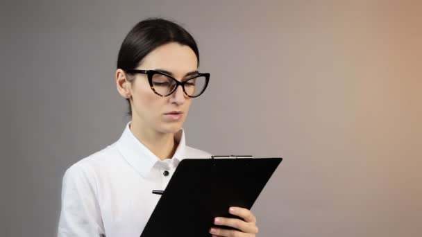 Trabajadora de oficina en gafas sosteniendo portapapeles sonriendo y toma notas sobre fondo gris en el estudio. Concepto de emociones — Vídeos de Stock