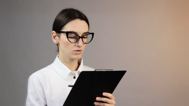 Travailleuse de bureau dans des lunettes tenant presse-papiers souriant et prend des notes sur fond gris au studio. Concept d'émotions — Video