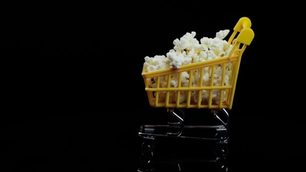 Gele winkelmand gevuld met popcorn wervelingen op een zwarte achtergrond ruimte voor tekst — Stockvideo