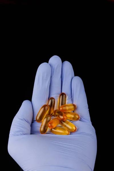 Omega-3 kapslar i hand med medicinsk handske. Hälsokoncept med fiskoljekapslar. På en svart bakgrund — Stockfoto
